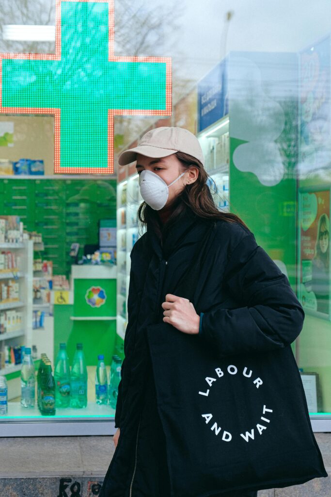 pexels-photo-3987219-3987219 Woman wearing a face mask carries a bag in front of a pharmacy.